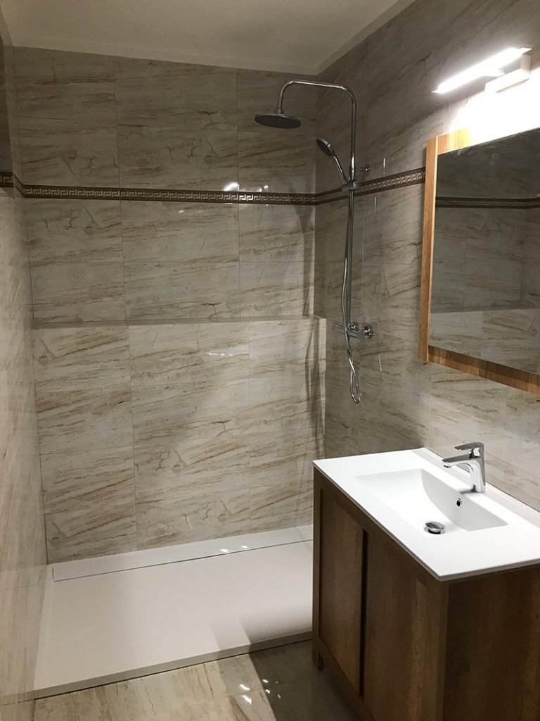 Ukończona łazienka z kabiną prysznicową typu walk-in, wykończona płytkami imitującymi marmur, z drewnianą szafką pod umywalką i lustrem w drewnianej ramie.