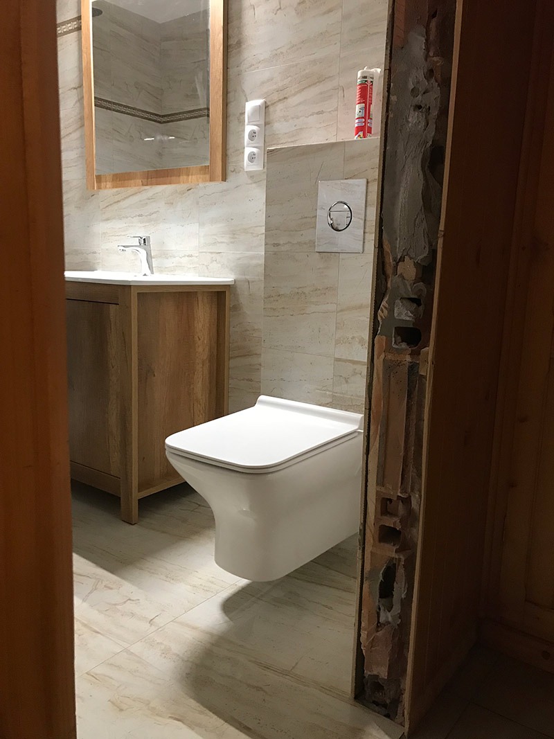 Nowoczesna toaleta podwieszana w trakcie montażu, widoczne fragmenty instalacji w ścianie, jasne płytki imitujące marmur na ścianach i podłodze, umywalka z szafką w tle.