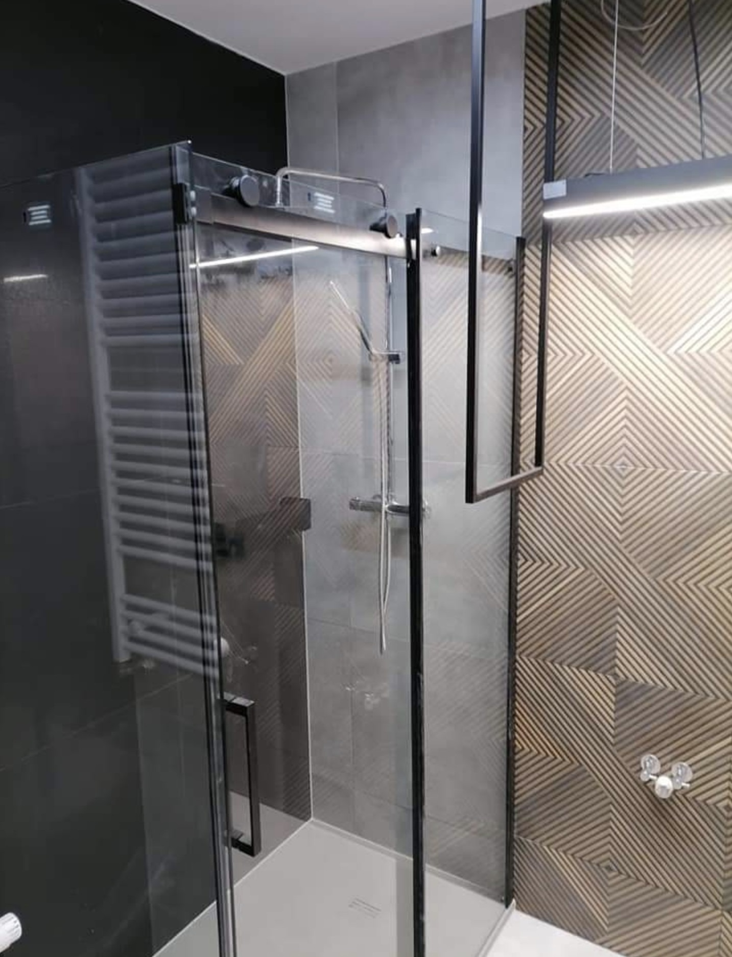 Nowoczesna kabina prysznicowa z czarnymi profilami i szklanymi drzwiami przesuwnymi, widoczna bateria prysznicowa i dekoracyjna płytka ścienna z geometrycznym wzorem.