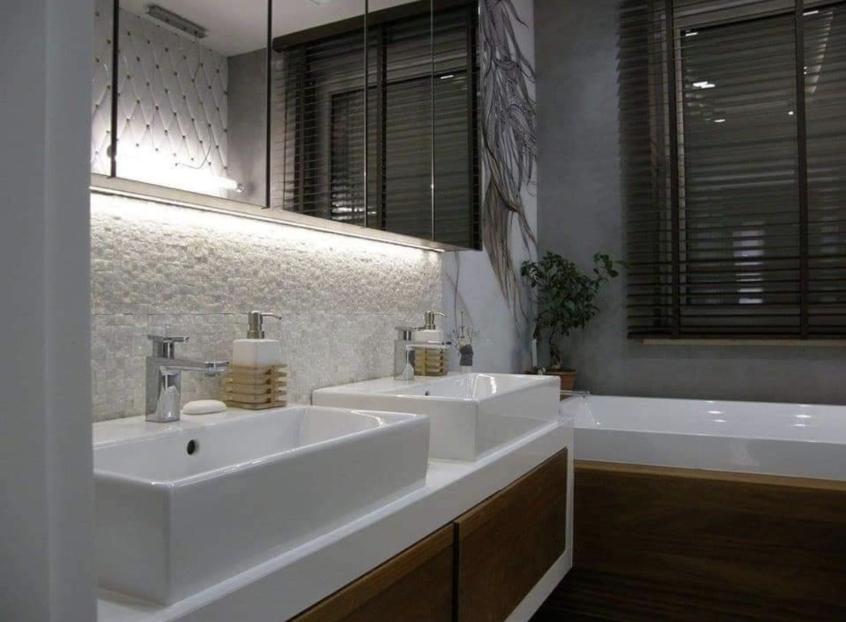 Nowoczesna łazienka z dwoma białymi umywalkami nablatowymi, chromowanymi bateriami, mozaiką na ścianie, podświetlanym lustrem, drewnianymi żaluzjami i dekoracyjnym rysunkiem na ścianie.