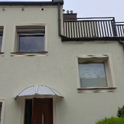 Jasna elewacja domu z dekoracyjnymi obramowaniami okien, brązowymi rynnami i balkonem z balustradą. Widoczne rolety zewnętrzne i markiza nad wejściem.