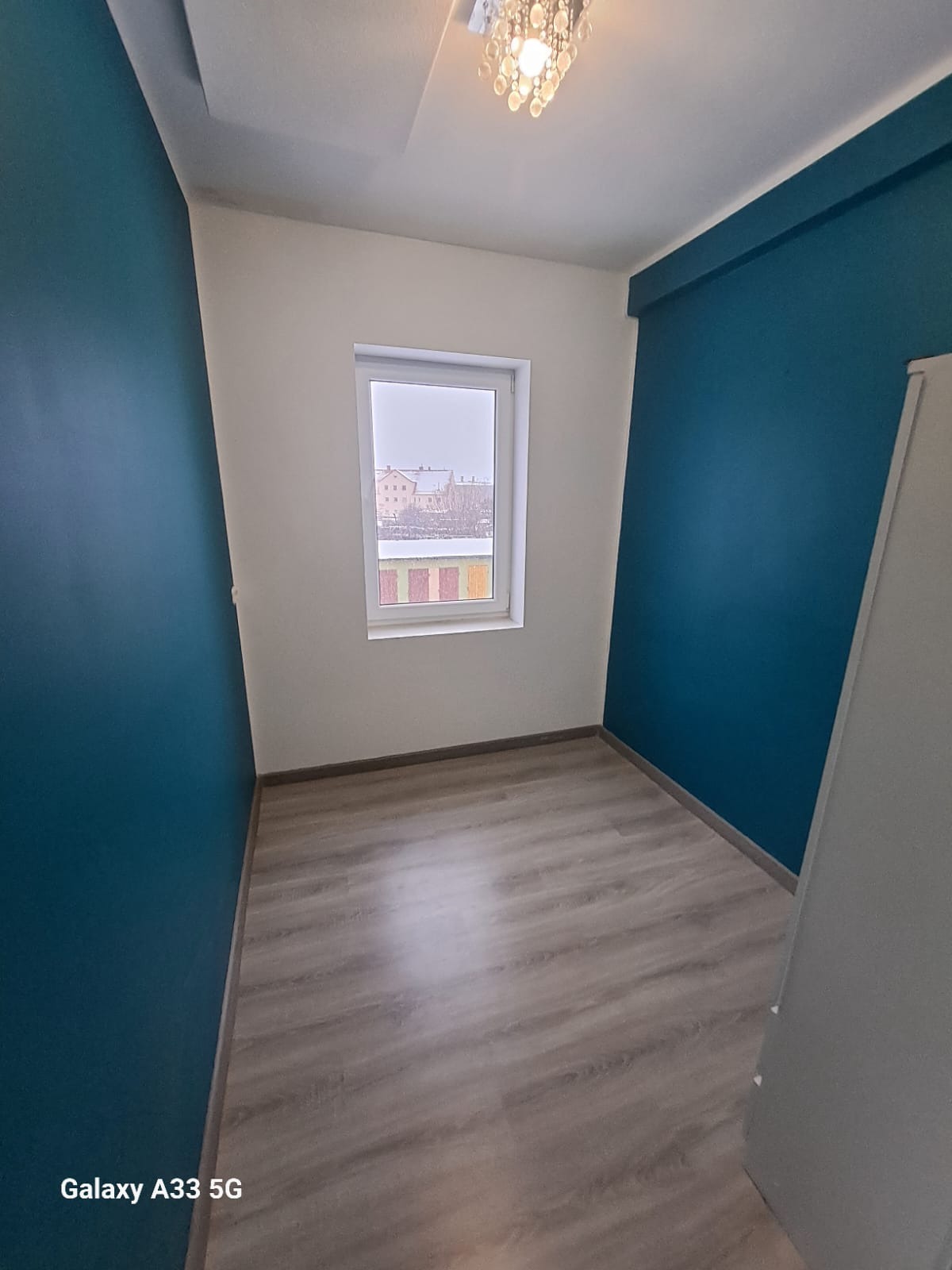 Pokój po remoncie: ściany pomalowane na biało i turkusowo, panele podłogowe ułożone, widok na okno z zimowym krajobrazem, żyrandol kryształowy na suficie.