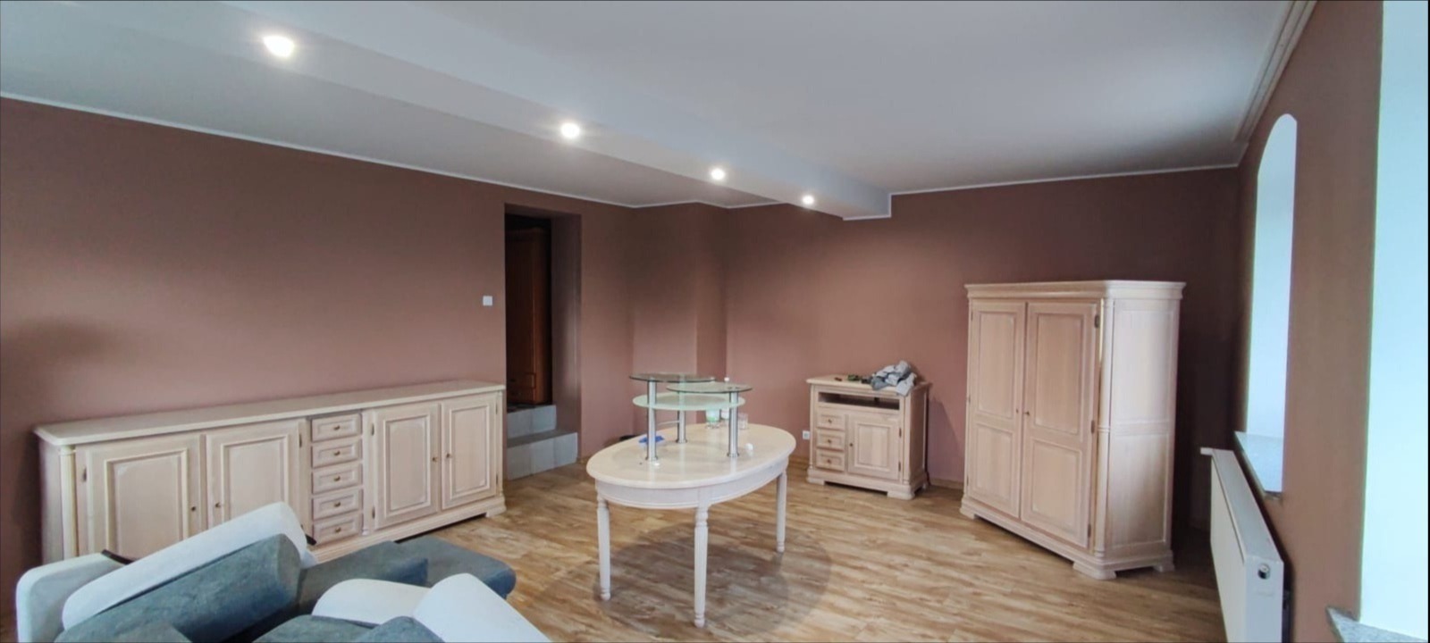 Pomieszczenie po malowaniu ścian na kolor brązowy, z jasnymi meblami w stylu retro i panelami podłogowymi, oświetlone punktowymi lampami sufitowymi.