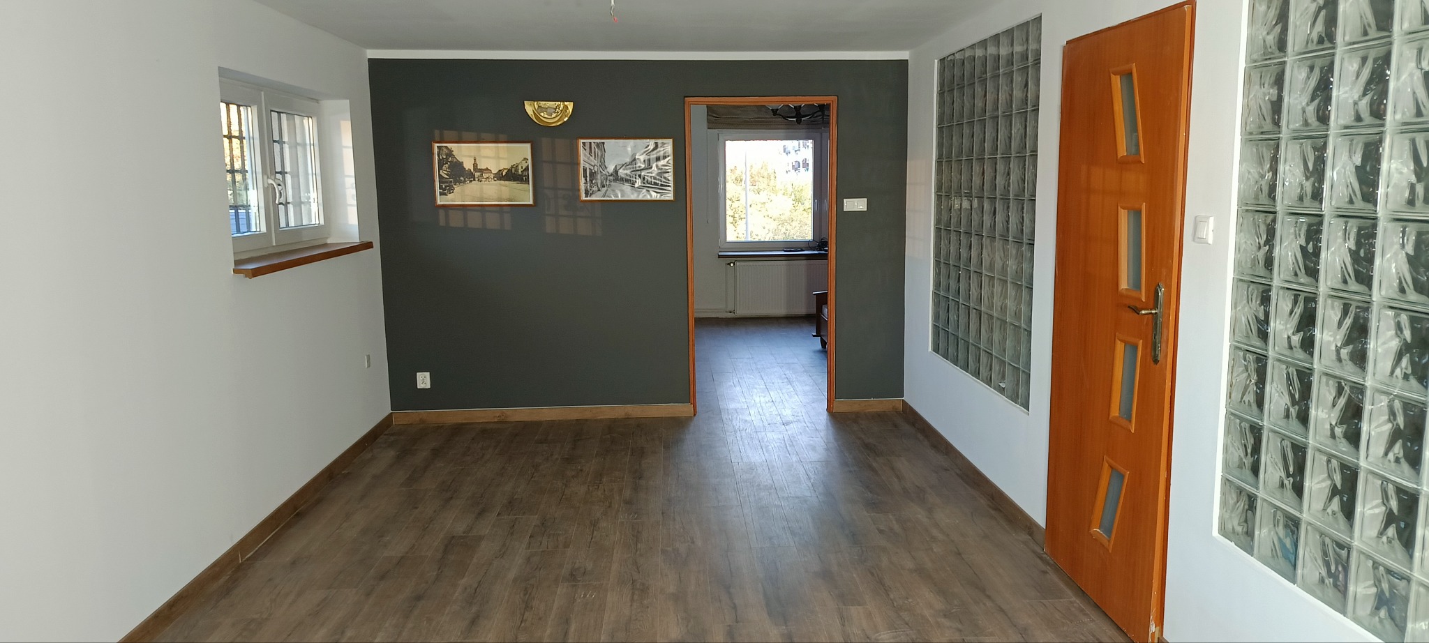 Pusta przestrzeń po remoncie z ciemnoszarą ścianą, drewnianą podłogą, oknem i drzwiami z luksferów, ukazująca efekt odświeżonego wnętrza.