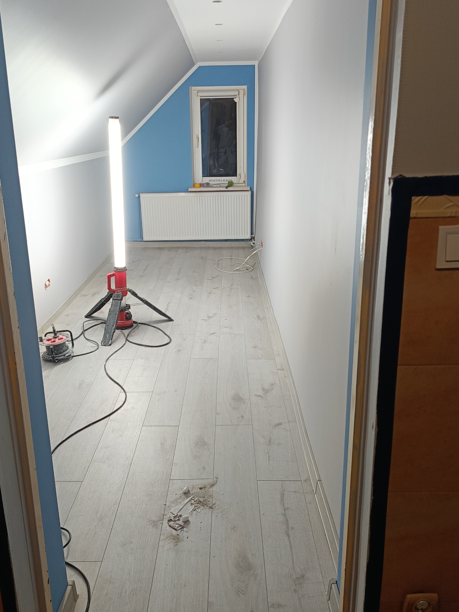 Wnętrze pokoju w trakcie prac wykończeniowych: ściany pomalowane na biało, fragment ściany bocznej na niebiesko, zamontowane panele podłogowe, widoczna lampa budowlana i resztki materiałów po...