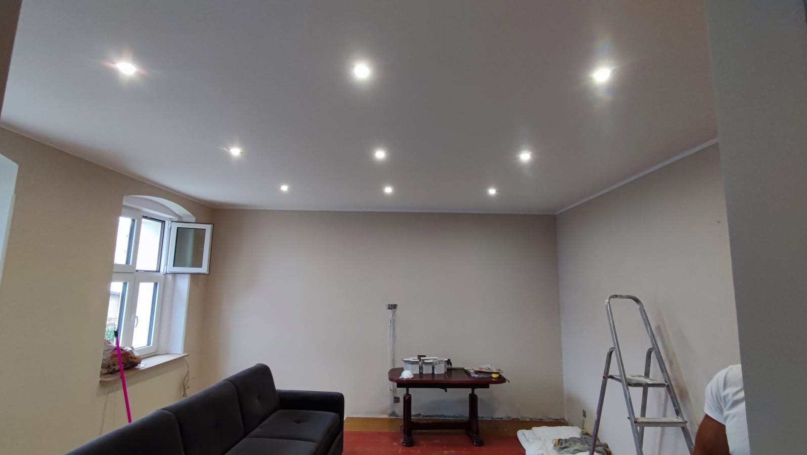 Wnętrze pokoju po malowaniu ścian na beżowo, z widocznym sufitem z oświetleniem punktowym, częściowo zasłoniętą sofą i stołem z akcesoriami malarskimi.