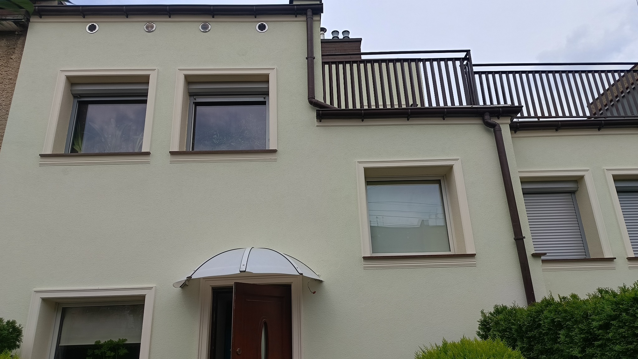 Jasna elewacja domu z dekoracyjnymi obramowaniami okien, brązowymi rynnami i balkonem z balustradą. Widoczne rolety zewnętrzne i markiza nad wejściem.