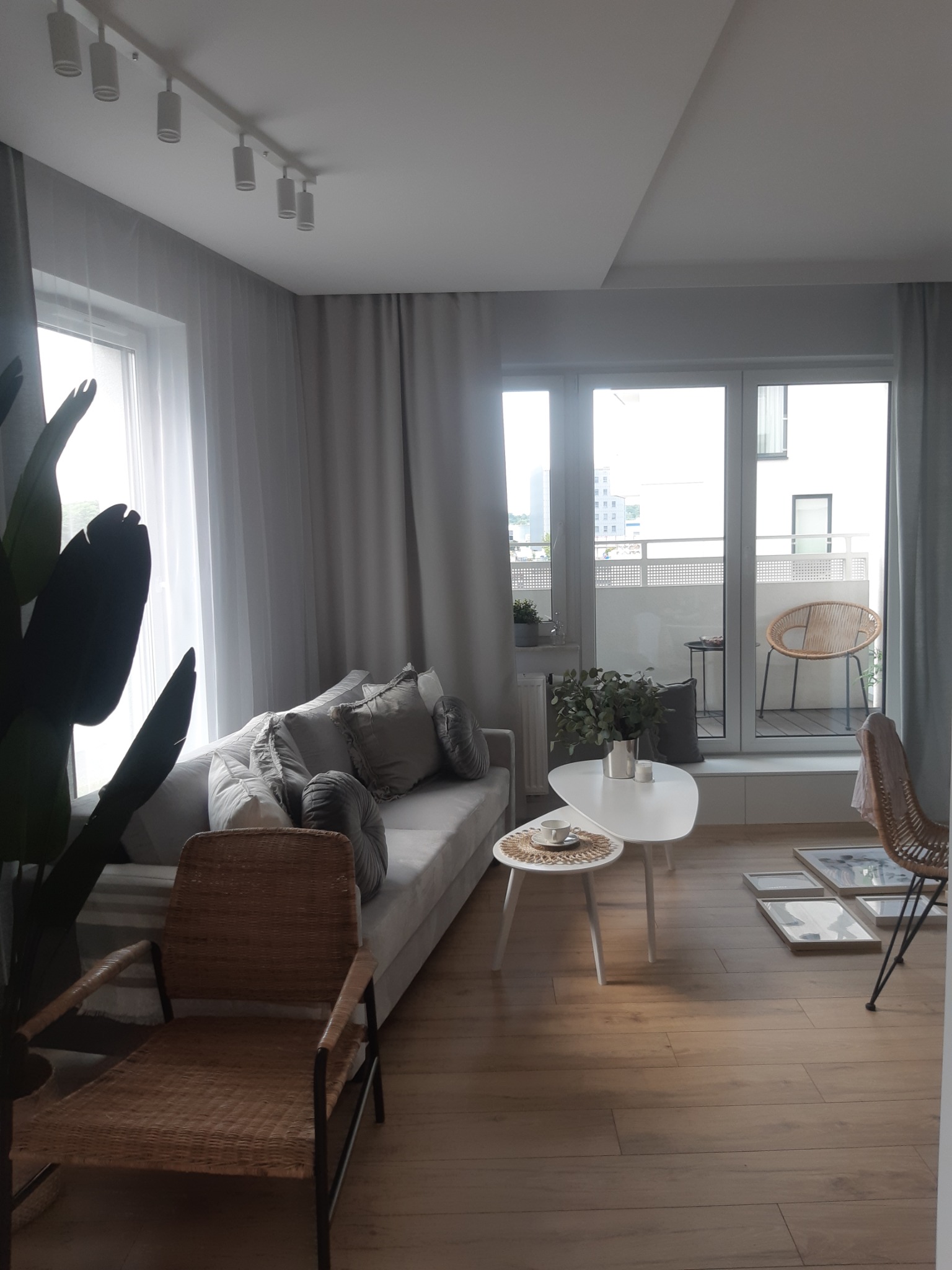 Jasne wnętrze salonu z sofą, wiklinowymi fotelami, stolikami kawowymi i dużym oknem balkonowym z widokiem na miasto.