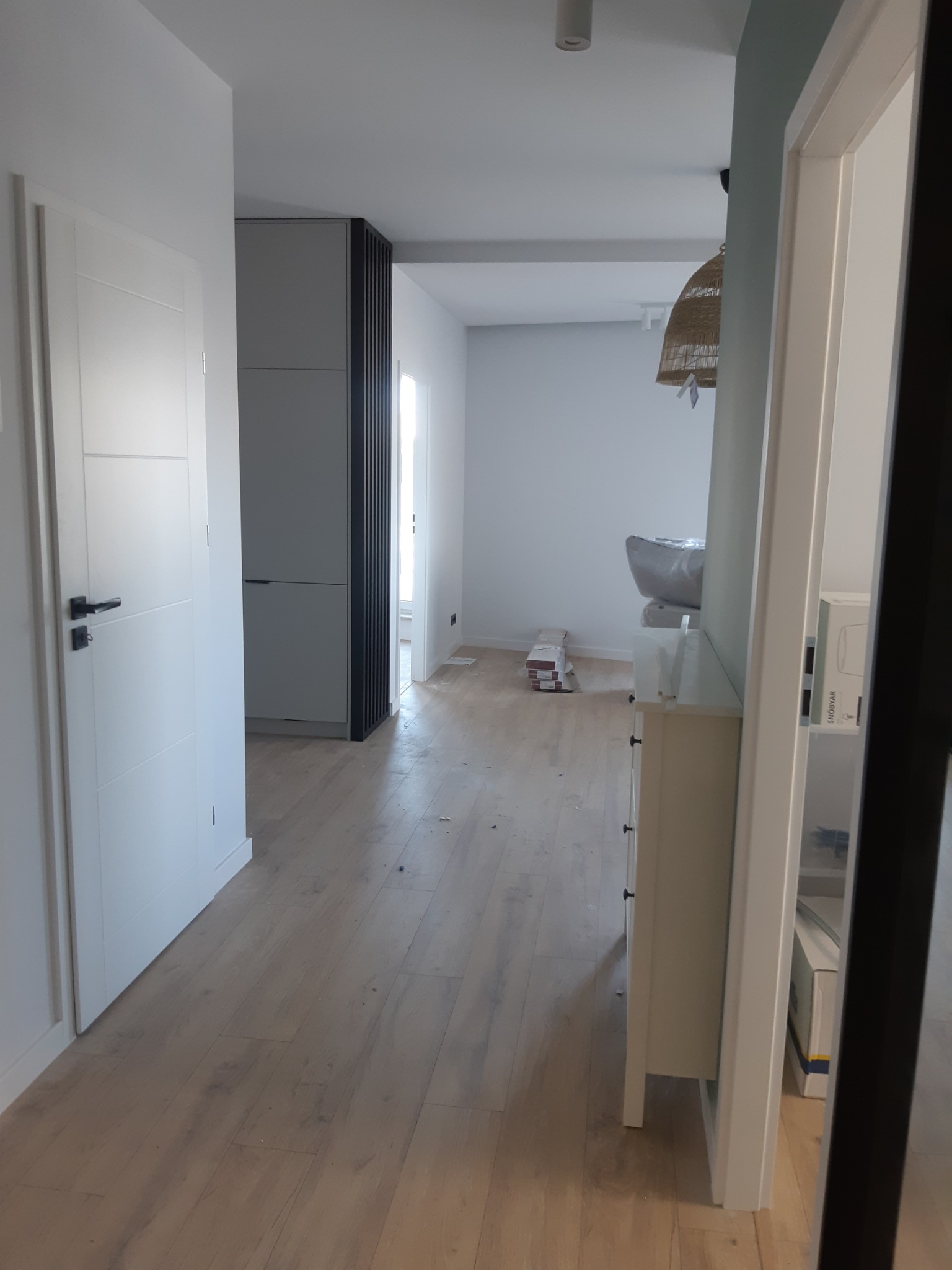 Korytarz w trakcie wykańczania z ułożoną drewnopodobną podłogą, widoczne białe drzwi z czarnymi klamkami, szara zabudowa meblowa oraz lampa z wikliny w tle.