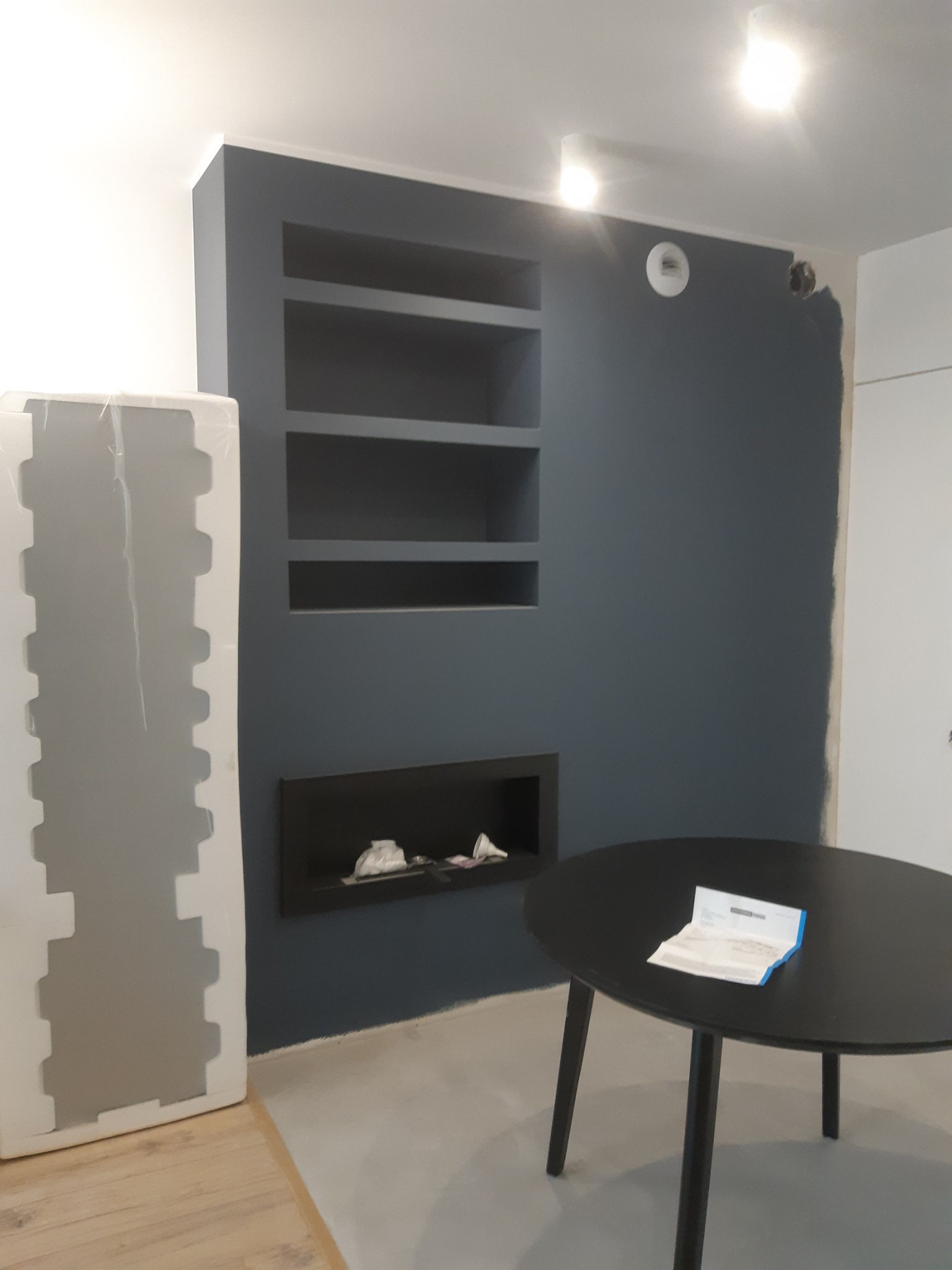 Niebieska ściana z wnękami i czarnym, wbudowanym biokominkiem, częściowo pomalowana, z podłogą w trakcie prac wykończeniowych i okrągłym stołem z dokumentami.