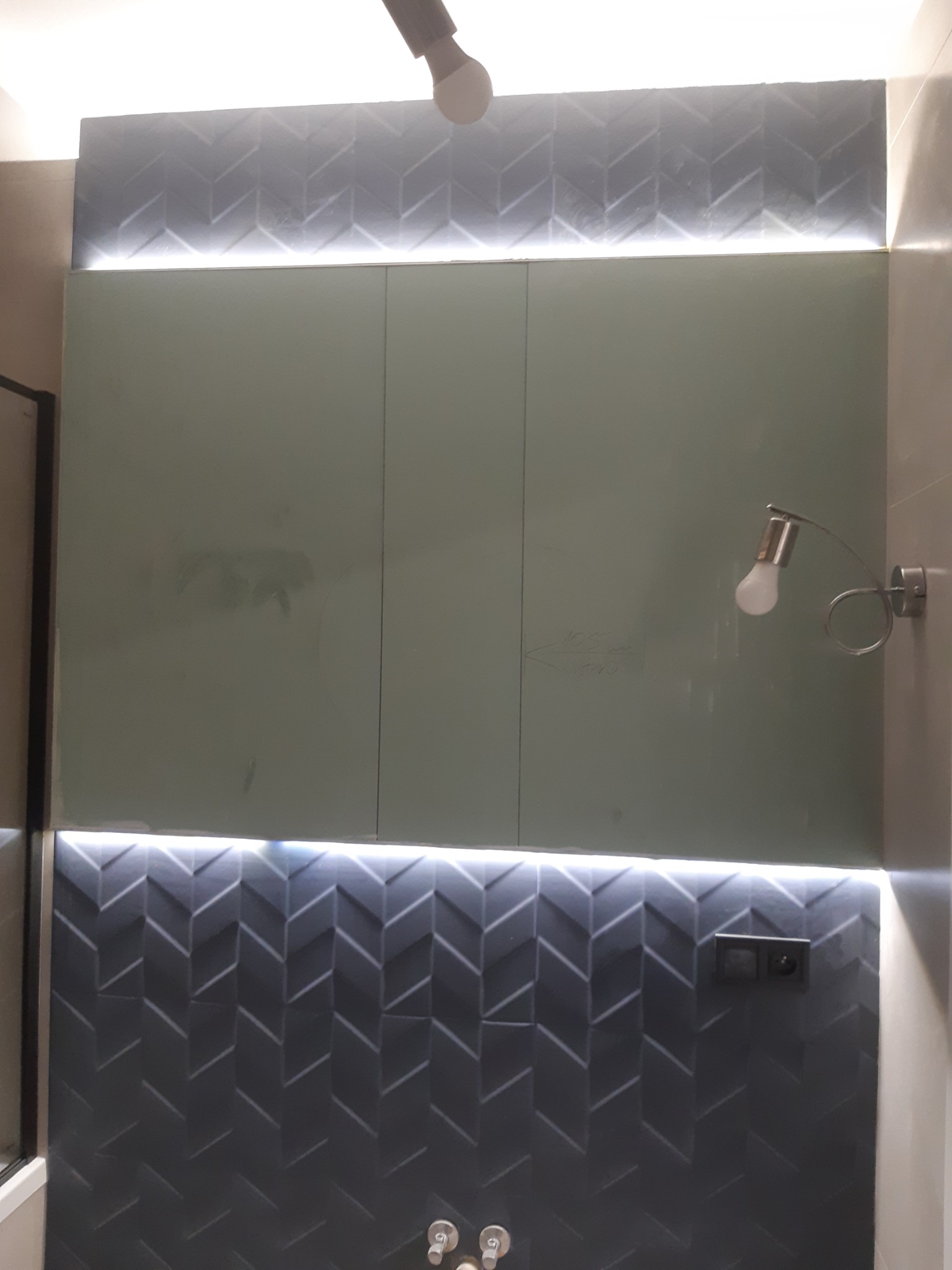 Nowoczesna łazienka z podświetlanymi panelami ściennymi w geometryczny wzór, jasnoszare szafki i lampa ścienna.