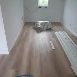 Pokój z nową podłogą z paneli, listwy przypodłogowe do montażu, zakryte folią przedmioty, widoczne gniazdka elektryczne.