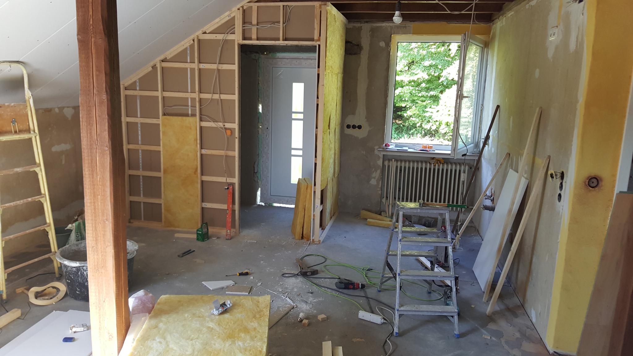 Wnętrze w trakcie remontu poddasza: drewniana konstrukcja szkieletowa ścian, izolacja z wełny mineralnej, widoczny fragment okna i grzejnika, narzędzia budowlane porozrzucane na podłodze, stara...