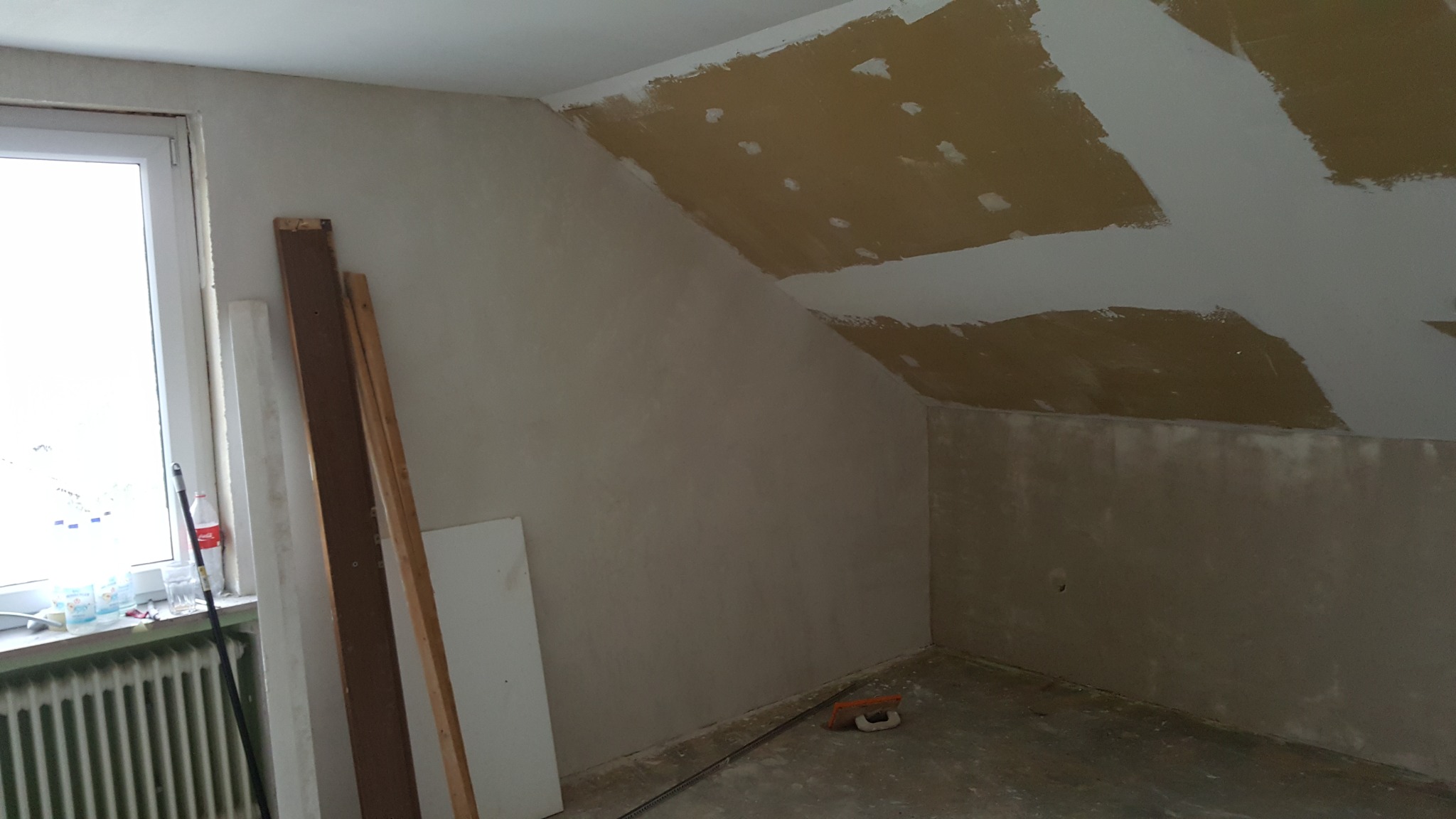 Pokój na poddaszu w trakcie remontu, ściany przygotowane do malowania, częściowo zagruntowane, widoczne płyty na suficie z nałożonym gruntem, narzędzia malarskie leżące na podłodze.