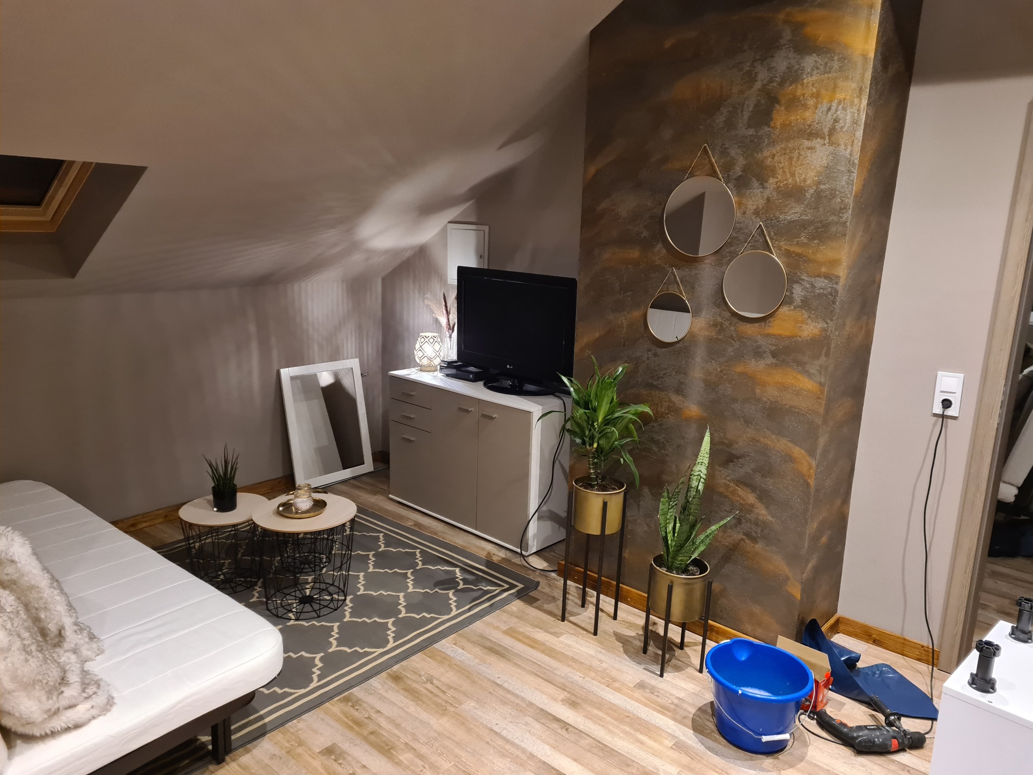 Pokój na poddaszu z dekoracyjną ścianą w odcieniach brązu i złota, trzema okrągłymi lustrami, roślinami doniczkowymi, sofą i narzędziami malarskimi w tle, sugerującymi prace wykończeniowe.