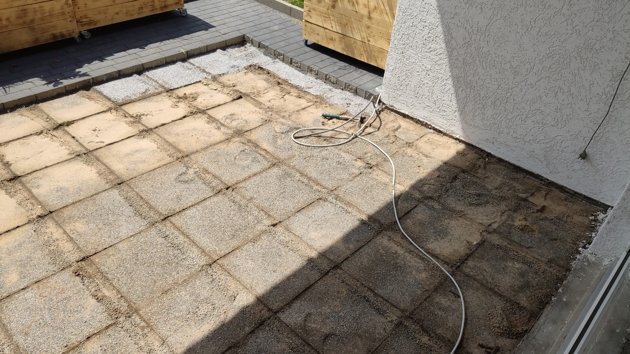 Niedokończona nawierzchnia z kostki brukowej, widoczne piasek i gruz, kabel elektryczny leżący na ziemi, fragment ściany budynku z fakturą.