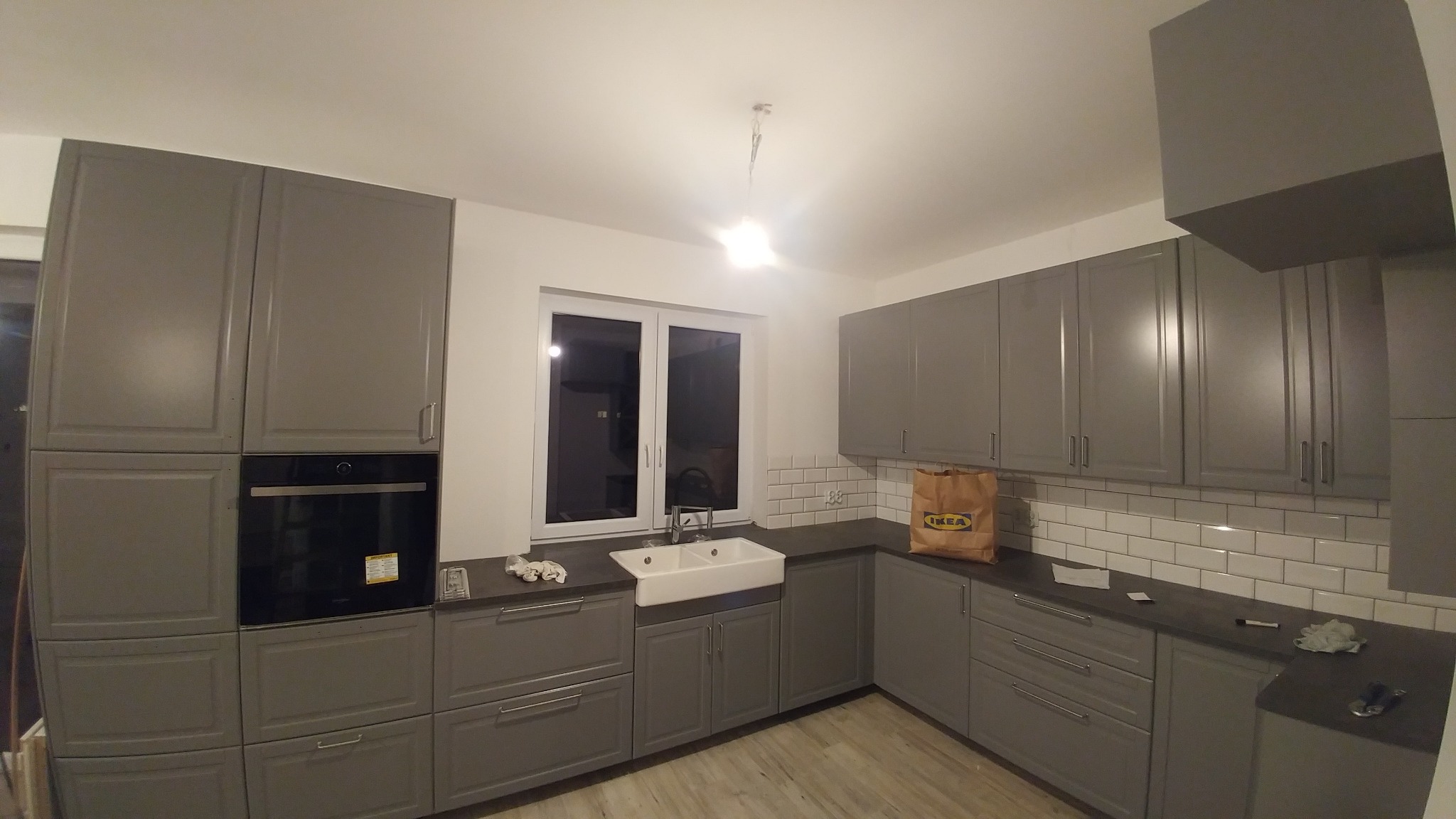 Szara kuchnia na wymiar z białym ceramicznym zlewem pod oknem, wykończona białymi płytkami typu metro, z widoczną torbą IKEA na blacie.