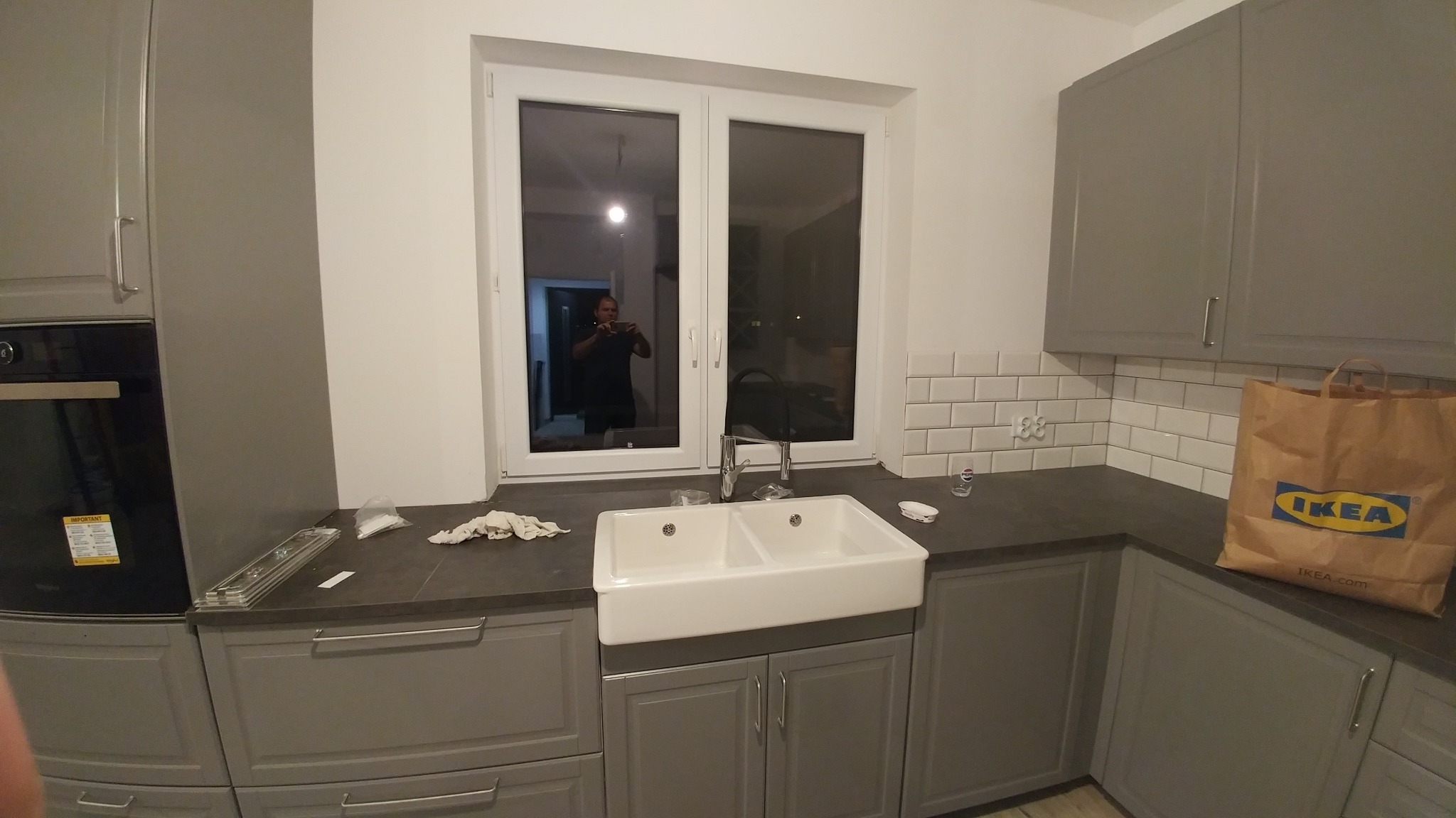Szare szafki kuchenne z białym ceramicznym zlewem dwukomorowym pod oknem, widoczna torba IKEA i odbicie osoby robiącej zdjęcie w oknie.