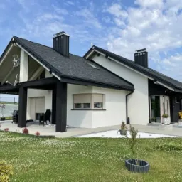 Nowoczesny dom jednorodzinny z białą elewacją i czarnymi elementami wykończeniowymi, widok z ogrodu na elewację frontową.