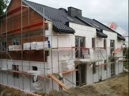 Budynek w trakcie prac termoizolacyjnych: widoczne rusztowania, częściowo ocieplona elewacja styropianem, niezabezpieczone otwory okienne, składowany materiał izolacyjny, dach pokryty czarną dachówką.