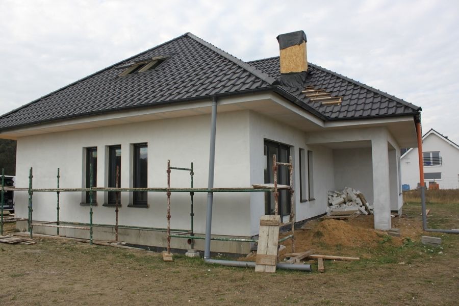 Nowoczesny dom jednorodzinny z białą elewacją w trakcie prac wykończeniowych, widoczne rusztowania, fragment odkrytej izolacji komina i sterta gruzu przy wejściu.