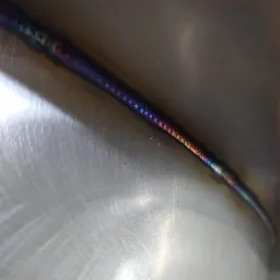 Szlifowana powierzchnia metalu ze świeżym, barwnym spawem TIG, widoczne tęczowe przebarwienia termiczne na spoinie.