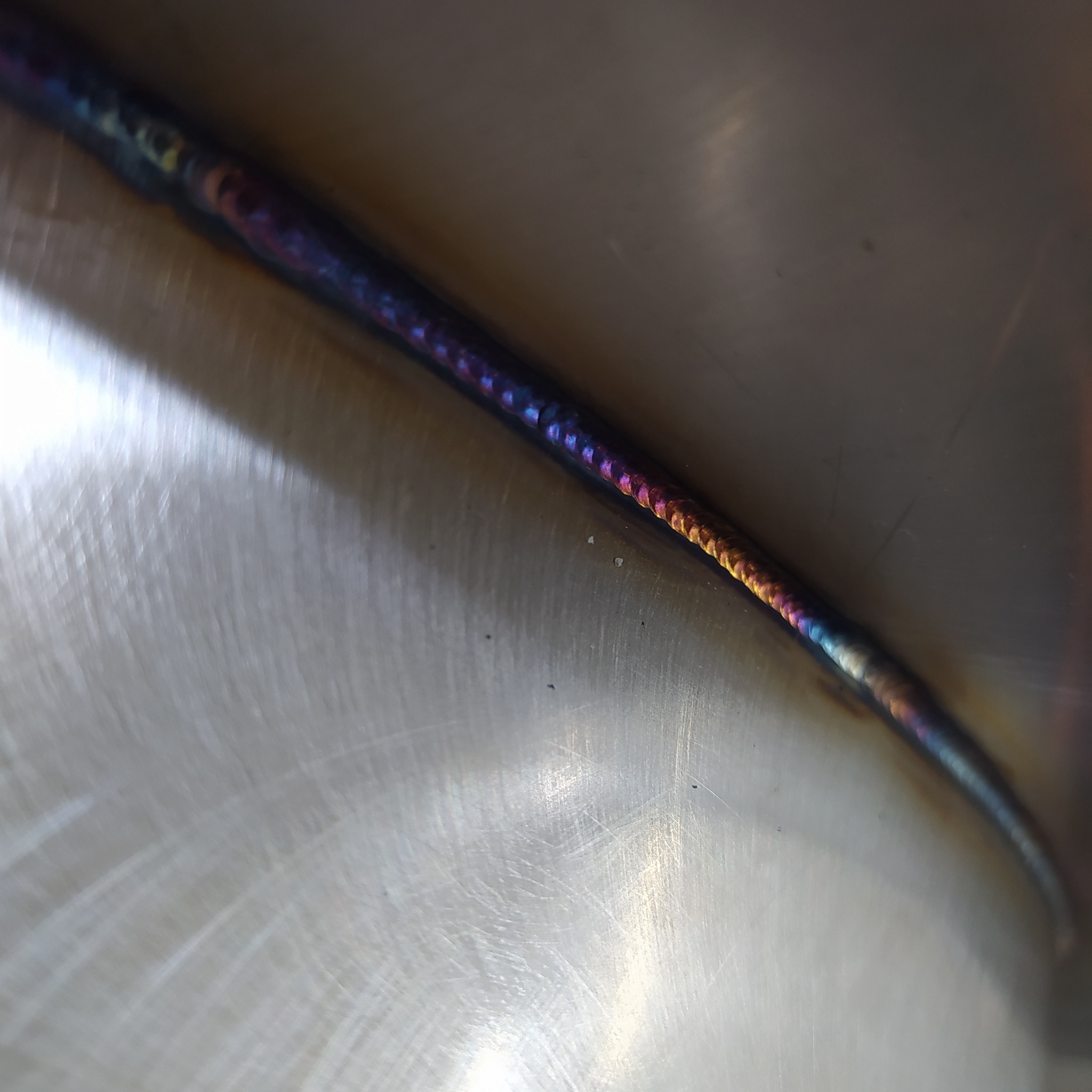 Szlifowana powierzchnia metalu ze świeżym, barwnym spawem TIG, widoczne tęczowe przebarwienia termiczne na spoinie.