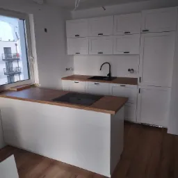 Nowoczesna, jasna kuchnia w trakcie montażu: białe szafki z czarnymi uchwytami, drewniany blat, czarny zlew i płyta indukcyjna na wyspie kuchennej, widoczne kable elektryczne zwisające z sufitu.