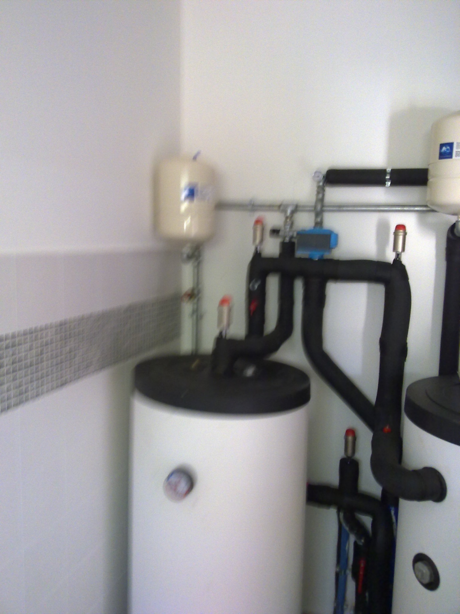 Białe zbiorniki akumulacyjne ciepłej wody użytkowej z czarnymi rurami w kotłowni, widoczne manometry i zawory bezpieczeństwa, instalacja w rogu pomieszczenia z szarą mozaiką na ścianie.