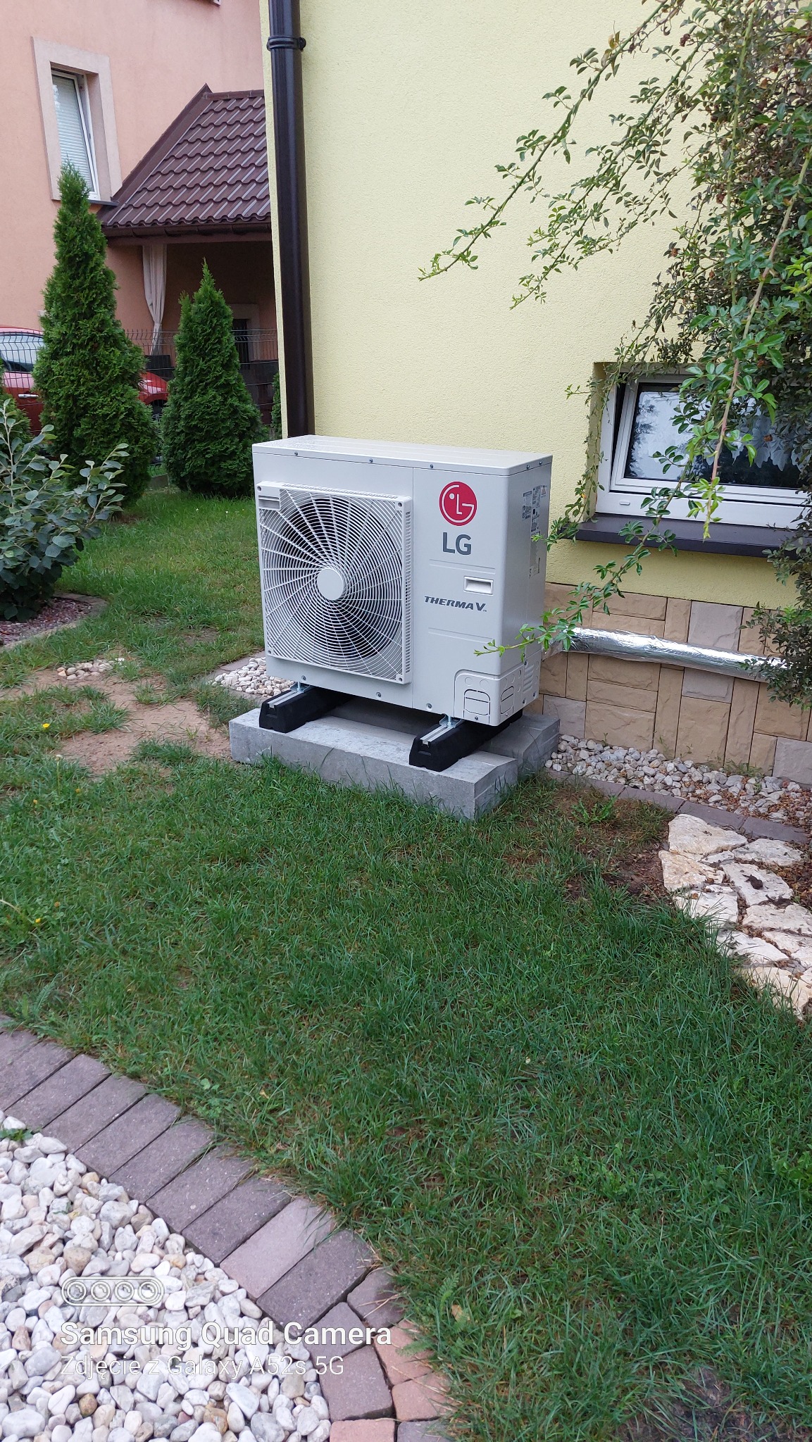Zewnętrzna jednostka klimatyzacyjna LG Therma V umieszczona na betonowej podstawie przed budynkiem z żółtą elewacją, otoczona trawnikiem i elementami dekoracyjnymi z kamienia.