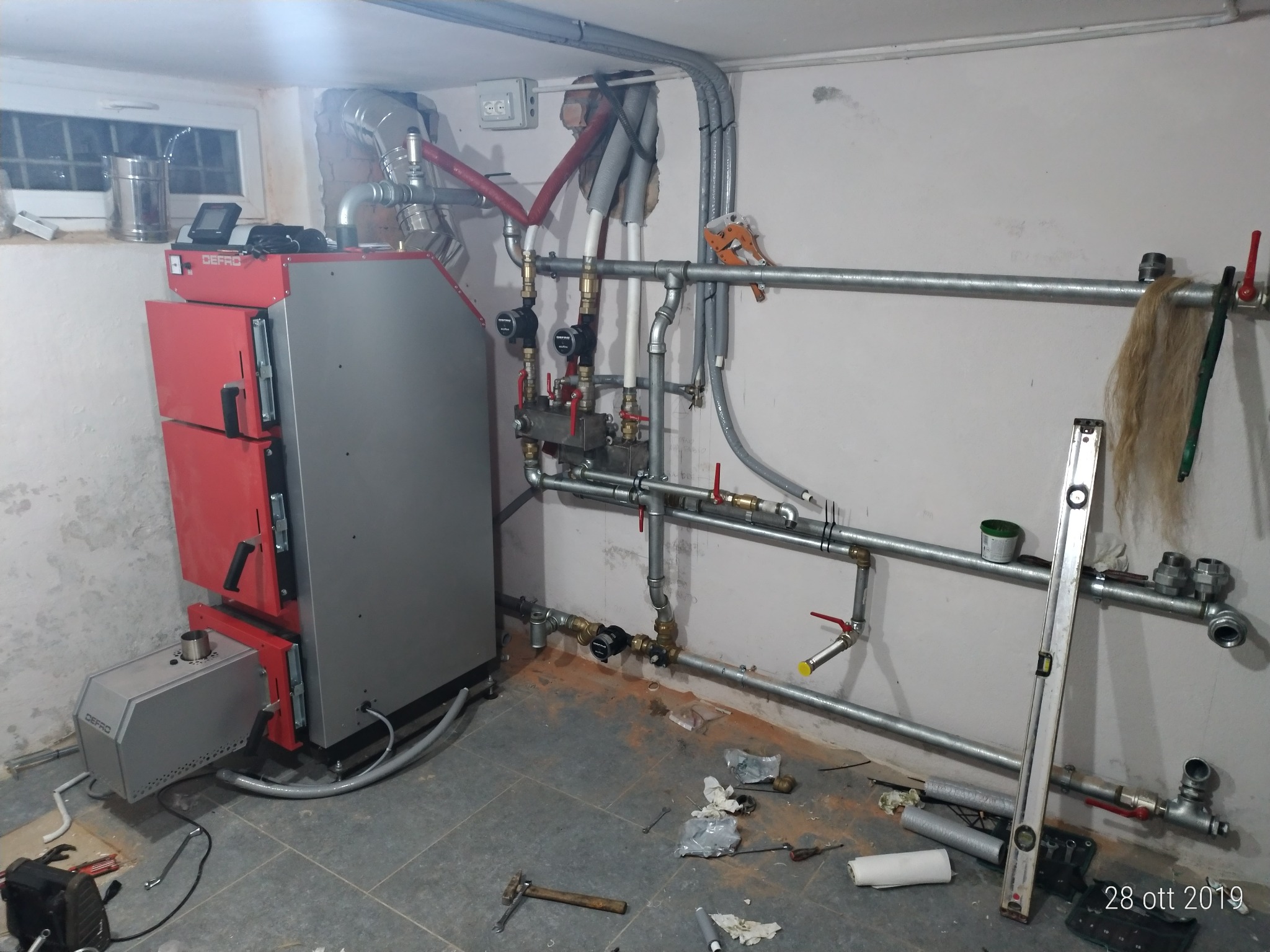 Instalacja kotła grzewczego DEFRo z podajnikiem, widoczne rury instalacji CO z zaworami, manometrami i pompami obiegowymi. Na podłodze narzędzia i materiały montażowe, poziomica oparta o ścianę...