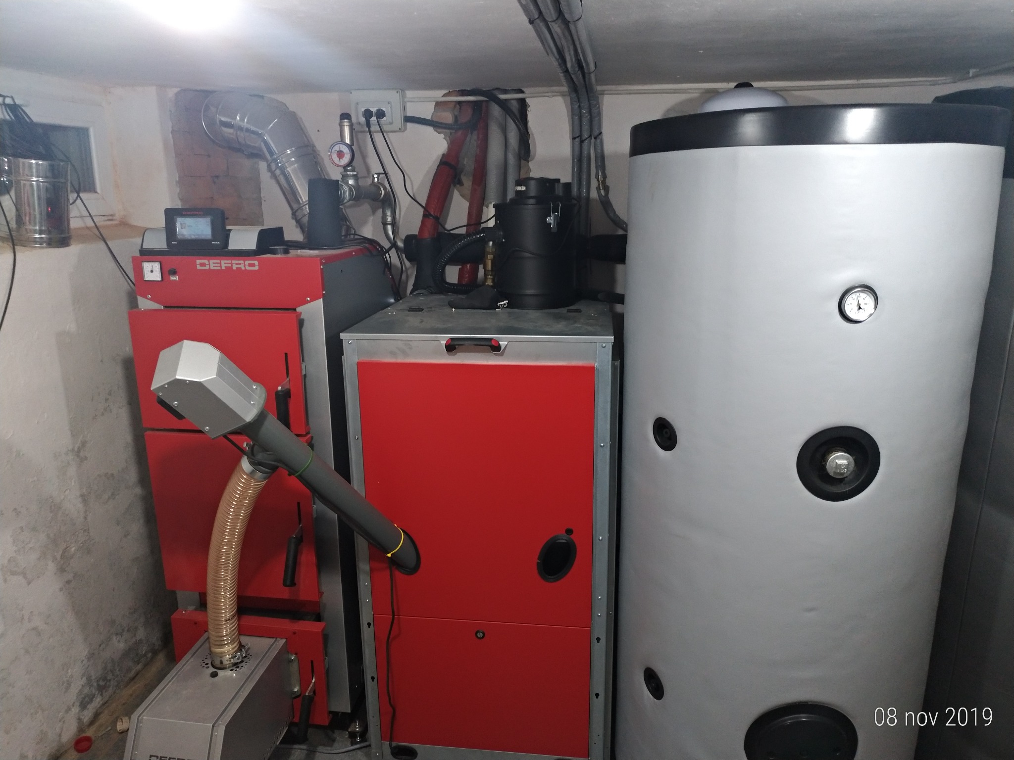Kotłownia z kotłem na paliwo stałe marki Defro, zasobnikiem ciepłej wody użytkowej i elementami instalacji grzewczej, widoczne przewody i osprzęt, pomieszczenie w piwnicy.