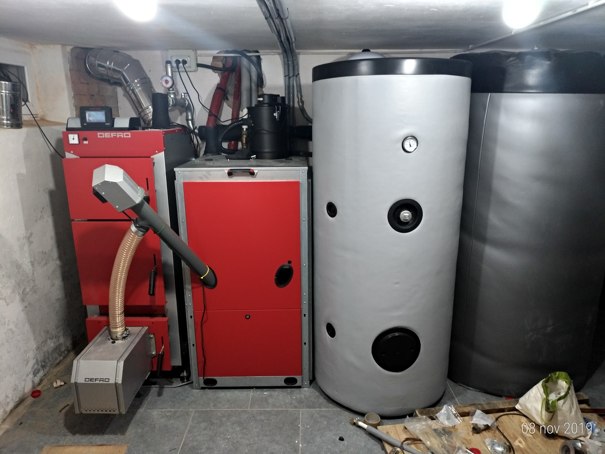 Czerwony piec z podajnikiem marki Defro, obok szary i czarny zbiornik akumulacyjny w kotłowni, widoczne elementy instalacji i narzędzia na podłodze.
