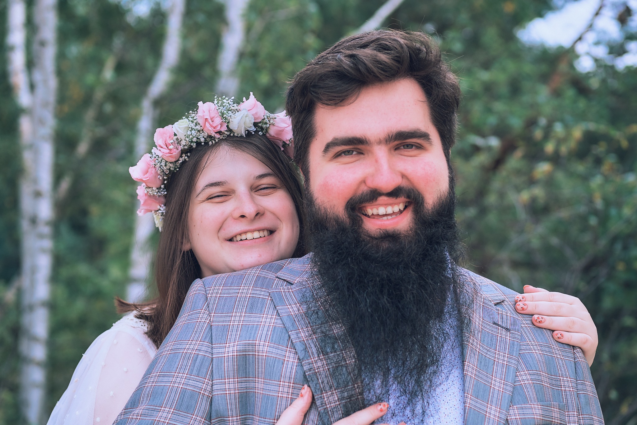 Uśmiechnięta para: kobieta w wianku z różowych kwiatów opiera głowę na ramieniu mężczyzny w kraciastej marynarce, oboje na tle drzew.
