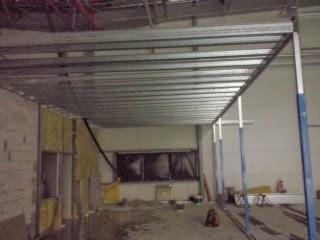 Stelaż sufitu podwieszanego z profili metalowych, widok z dołu, surowy stan, instalacje widoczne w tle, fragment ściany z żółtą izolacją.