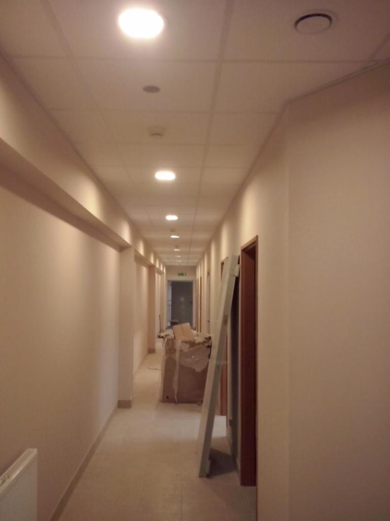 Długi korytarz biurowy z sufitem kasetonowym i wbudowanym oświetleniem. Widoczne przygotowania do prac wykończeniowych: drzwi oparta o ścianę, zabezpieczone materiały.