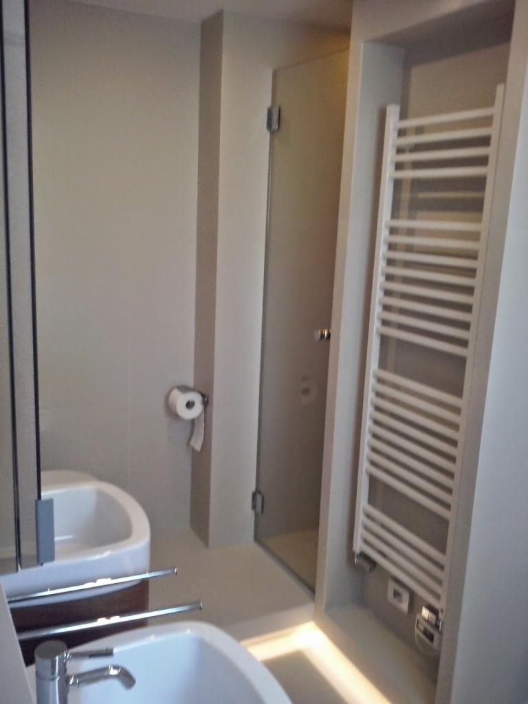 Nowoczesna łazienka w beżowej tonacji z grzejnikiem drabinkowym, kabiną prysznicową ze szklanymi drzwiami i umywalką. Widoczny dozownik papieru toaletowego i oświetlenie LED.