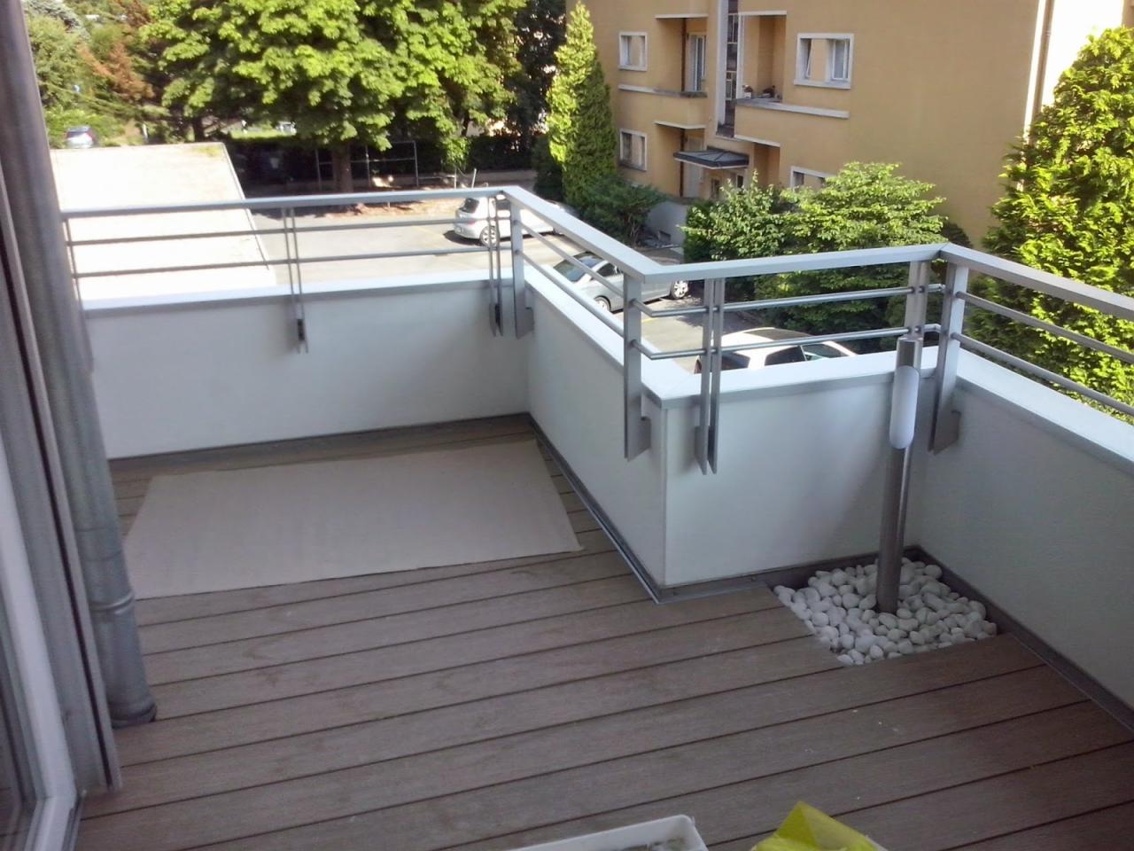 Wykończony balkon z drewnianą podłogą, białymi ścianami i metalową balustradą. Dekoracja z białych kamieni przy słupku oświetleniowym. Widok na zieleń i budynek.