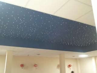 Niebieski sufit z efektem gwiazd w jasnym wnętrzu. Dekoracyjne oświetlenie punktowe tworzy unikalny klimat. Widoczne trzy czerwone dekoracje na ścianie.