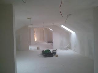 Poddasze po gipsowaniu ścian i sufitu, widoczne przewody elektryczne, narzędzia malarskie na podłodze, okno dachowe w skosie. Wnętrze w odcieniach bieli.