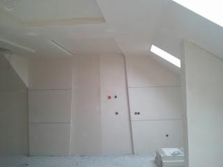 Poddasze po montażu płyt gipsowo-kartonowych. Widoczne łączenia płyt i otwory na gniazdka. Ukośny sufit z oknem dachowym. Prace wykończeniowe w jasnym wnętrzu.