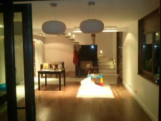 Nowoczesny salon po remoncie: drewniana podłoga, białe ściany, minimalistyczny kominek, schody z białymi stopniami i designerskie lampy wiszące.