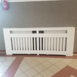 Biała, drewniana obudowa grzejnika z pionowymi szczebelkami, wykonana w Bolkowie, na tle szarej ściany i podłogi z płytek w odcieniach beżu i brązu. Widoczna gaśnica na ścianie.