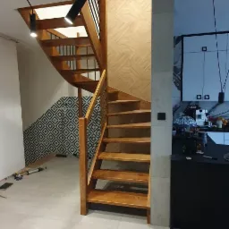 Kręte, drewniane schody z metalową balustradą w nowoczesnym wnętrzu z geometrycznymi wzorami na ścianie i podłodze.