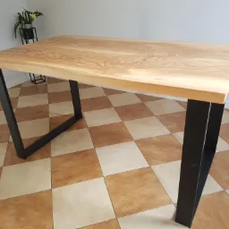 Stół z blatem z jasnego drewna o wyraźnym usłojeniu, na czarnych, metalowych nogach w kształcie trapezu, na tle kafelkowej podłogi i ściany z rośliną w tle.