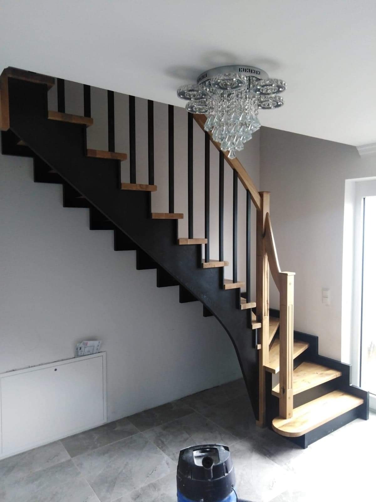 Nowoczesne schody z drewnianymi stopniami i czarną metalową konstrukcją, oświetlone kryształowym żyrandolem, z widocznym odkurzaczem na podłodze.