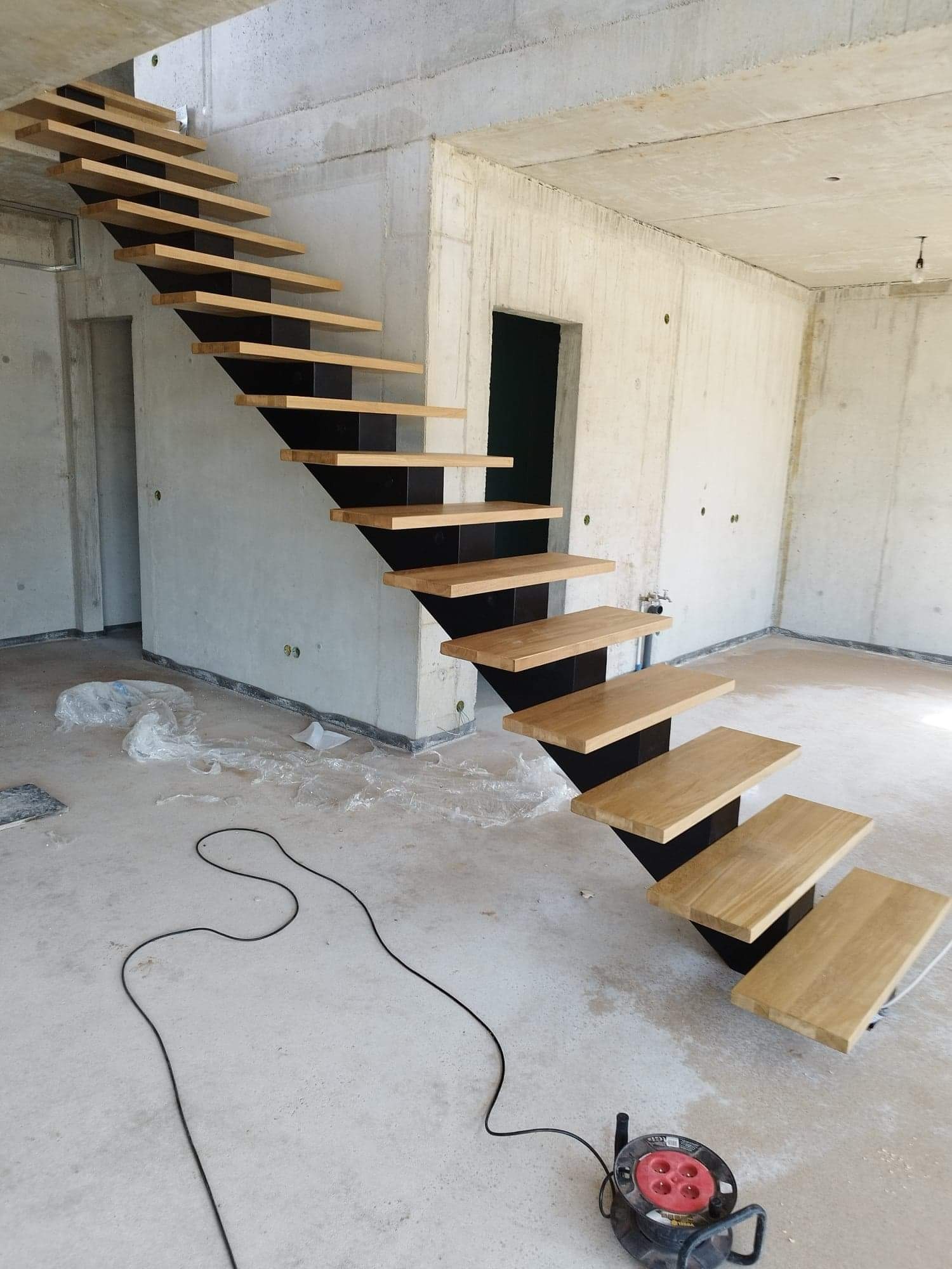 Surowe wnętrze z betonowymi ścianami i zawieszonymi drewnianymi stopniami schodów na czarnej, metalowej konstrukcji, widoczny zwój kabla elektrycznego na podłodze.