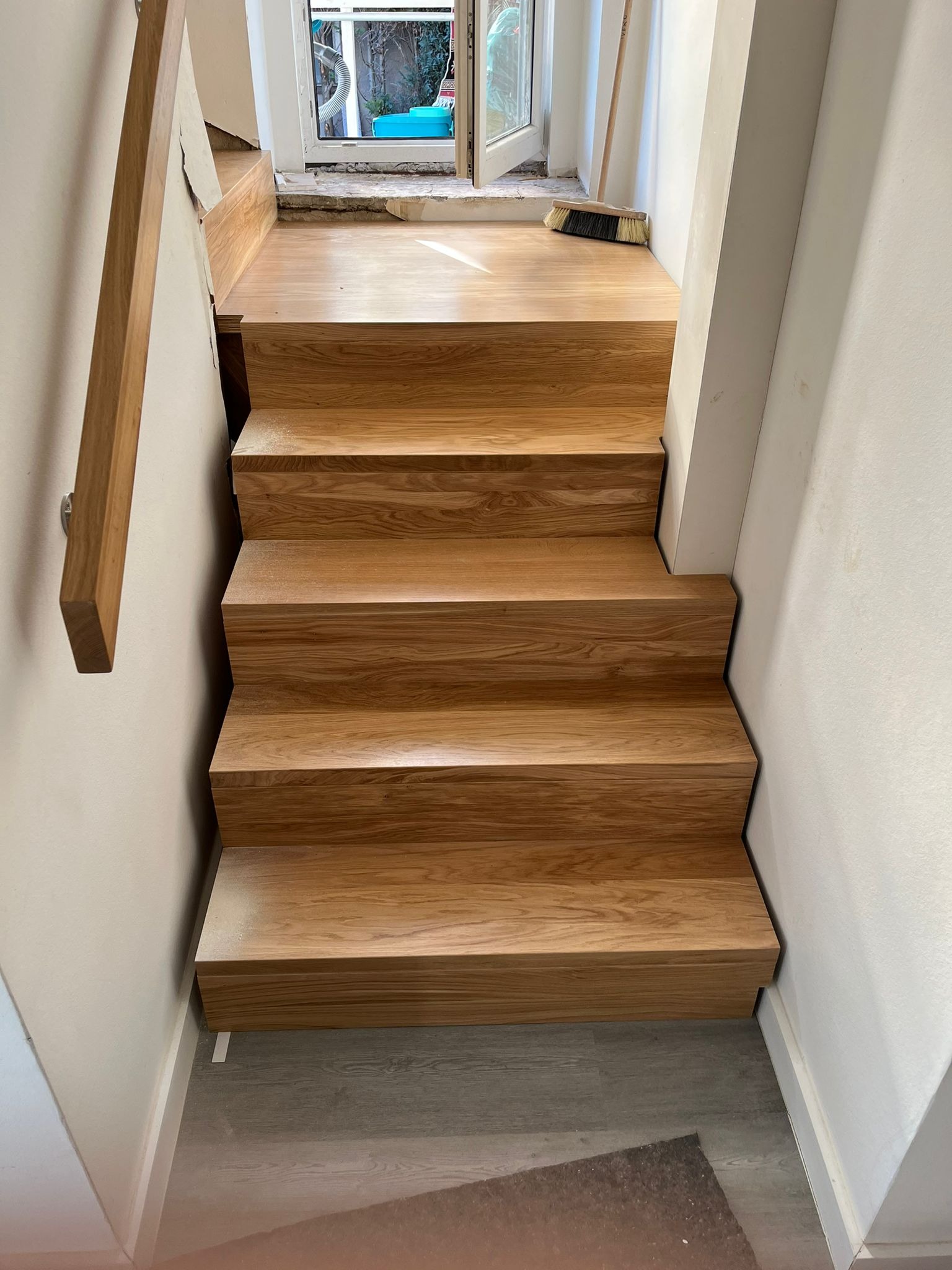 Wewnętrzne schody drewniane z poręczą, częściowo wykończone, z widocznym oknem i szczotką na górnym stopniu, w trakcie prac wykończeniowych.