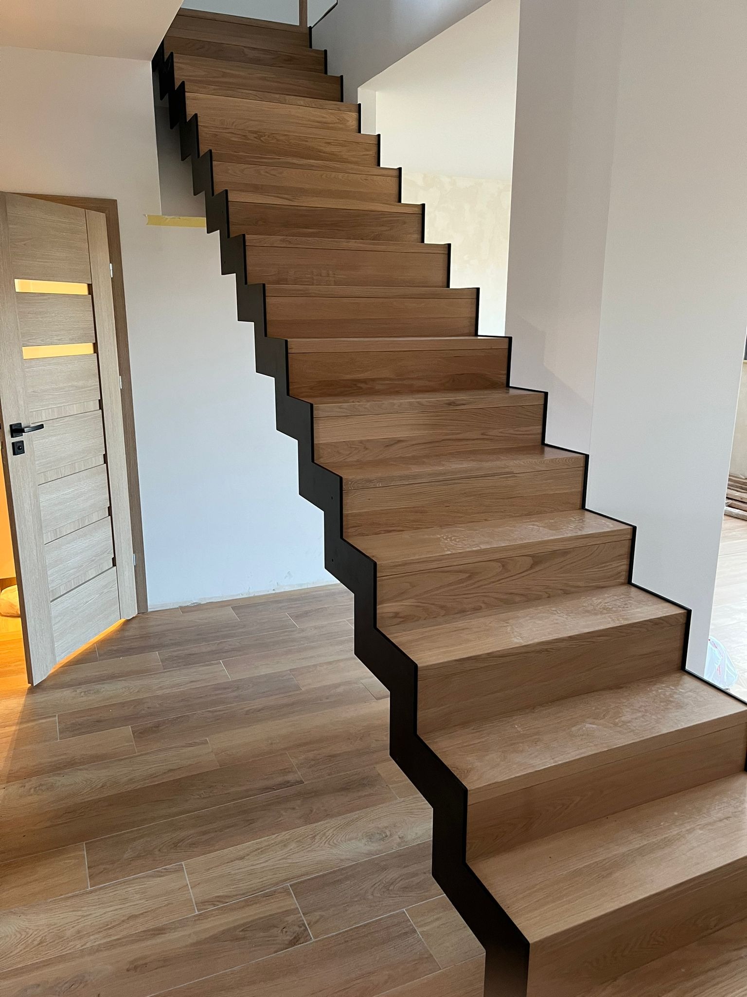 Nowoczesne, dębowe stopnie z czarnymi podstopnicami, tworzące schody w Bolkowie. Widoczna drewniana podłoga i drzwi z przeszkleniem.