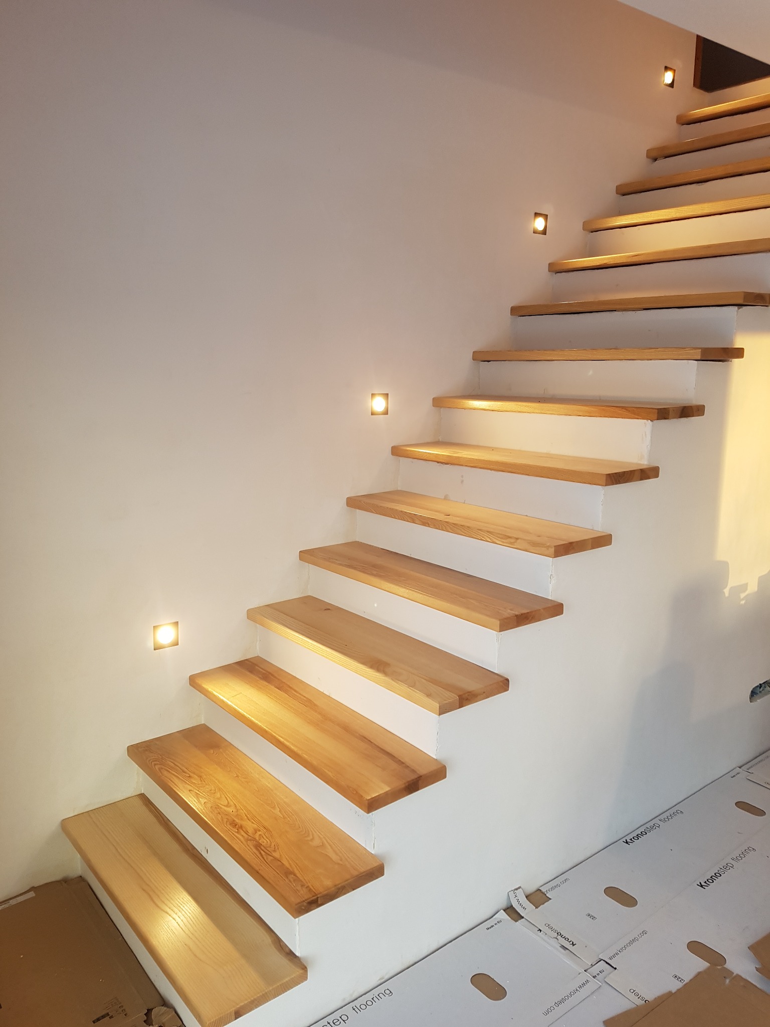 Nowoczesne, jednobelkowe schody z jasnego drewna z podświetleniem punktowym na białej ścianie, widoczne zabezpieczenia podłogi podczas montażu.